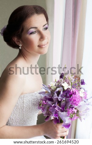 Bride waiting for the groom. She stands at the window, smiling and looking into the distance. In the hands of her purple wedding bouquet.