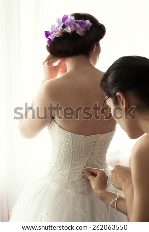 Bride in wedding dress standing at the window. On the dress laced corset. Help the bride get dressed. Corset laces on a wedding dress. Iris flowers in her hair.