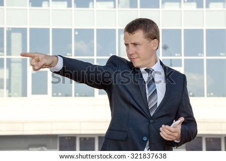 Business man on the background of an office building. He points his finger to the side. Pointing to the side. In his hand phone.