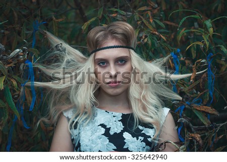 Young girl with blond hair and mystery look stands near an autumn tree. The girl look like a witch. Her hair is tied to branches of the tree and it seems that they levitate. Photo with cold toning.
