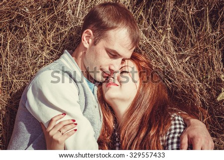 Young couple: attractive man and woman with red hair lying on a stack of hay, relaxing, kissing, embracing and enjoying the autumn sun. Photo with warm toning