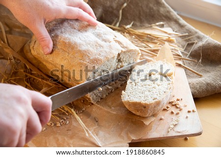 Woman slices fresh bread. hands with a knife. Freshly baked rye bread on a wooden board. Burlap, craft paper and straw on the background. Organic bread baked at home from wheat flour. Sliced bread.