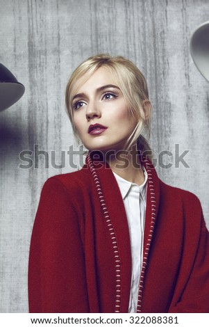 Closeup of beautiful young blond woman wearing long red coat.\
Her hair is tied into fashion ponytail and fresh natural make up suit to the style. Composition is fill with concrete wall and red mailbox