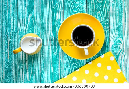 cup of black tea on yellow plate and yellow milk jug\
\
on turquoise colored old wooden table with yellow napkin at polka dots top view