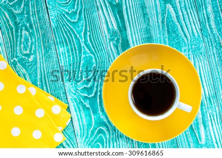 cup of black tea on yellow plate and yellow milk jug on turquoise colored old wooden table with yellow napkin at polka dots top view