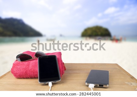 The concept idea. Red tower, sun glasses, mobile charging with power bank over wooden table on blur beach sand and blue sky background.