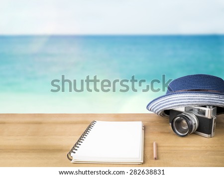 Idea concept. Empty notebook page, blue hat and old vintage camera over wooden table on sunshine blue sky and ocean background.