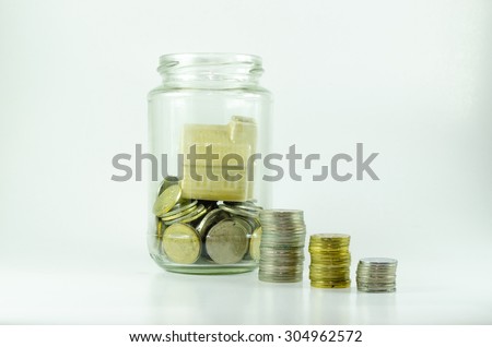 Miniature house inside open glass jar filled with coins, stacked coins outside isolated on white background.