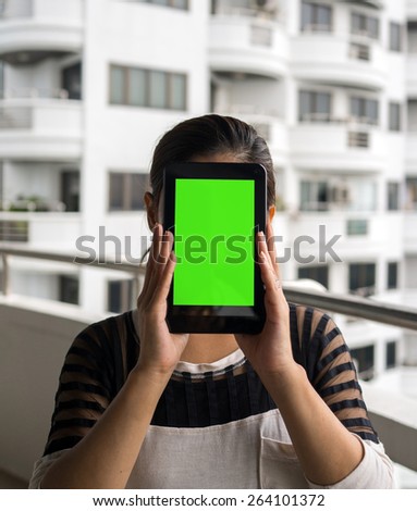 Asian woman holding tablet in her hands in front of her face with blank green screen and white building in the background.