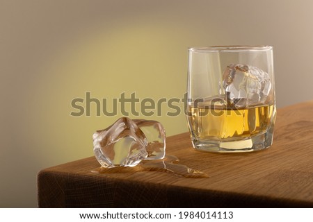 Whiskey or alcoholic beverage, with ie in a square formed designer glass. Sitting on an Oak table, with a backlighted gray surface. With melting Ice cubes on the side of the glass.  Imagine de stoc © 