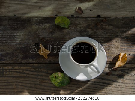 Cup of coffee , dried leaves,and green leaves on wooden table under the sun and shadows