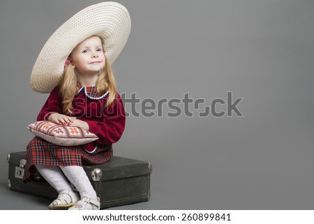 Happy And Smiling Caucasian Child Posing in Big Round Sombrero Hat and Sitting on Outdated Suitcase. Against Gray Background. Horizontal Image