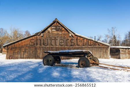 Horse carriage with car wheels and shafts that lie ahead in the snow, on a background rural wooden barn. Tomcats lie on the projections of the barn and bask in the bright low winter-spring sun