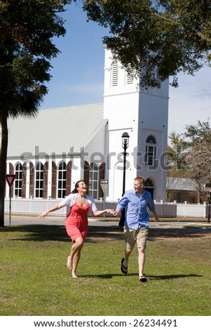An engaged couple hold hands as they have fun, running by a church.