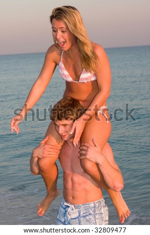 A young man gives a bikini clad woman a piggyback ride at sunset on the beach.