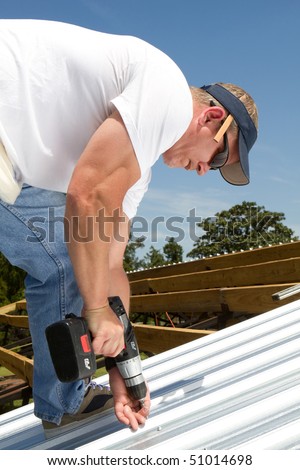 Roofer construction worker uses a battery powered screw-gun to fasten sheets of metal roofing to the rafters on top of a barn.