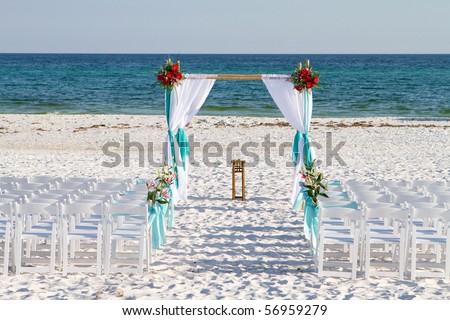 Wedding archway, chairs and flowers are arranged on the sand in preparation for a beach wedding ceremony.