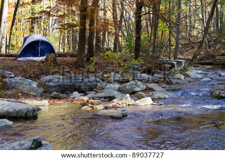 Small camping tent is pitched by a mountain stream in the woods in autumn with fall foliage.