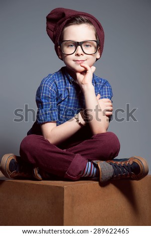 A blue - eyed kid with glasses. A boy is sitting with so merry face. He is wearing plaid shirt and burgundy pants with hat