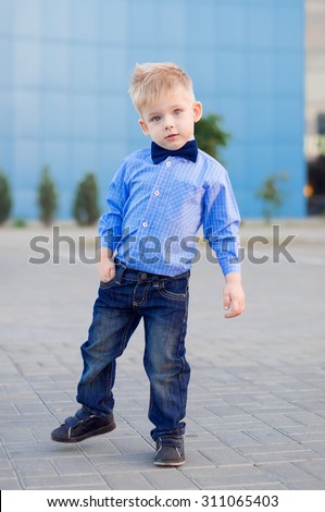 Stylish baby boy with blond hair. A boy is wearing the checked shirt, jeans and blue bow tie . He poses near a shopping center. Outdoor shot