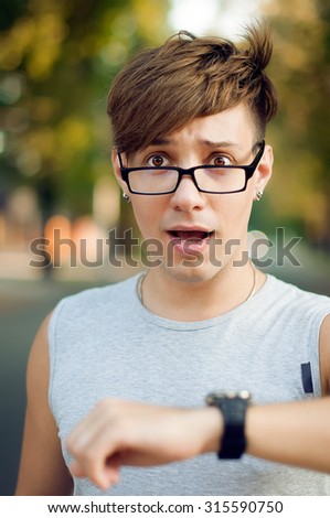 Attractive young man looking at his wristwatch in autumn park. He is wearing glasses. Hipster style. Sunny weather. Outdoor shot