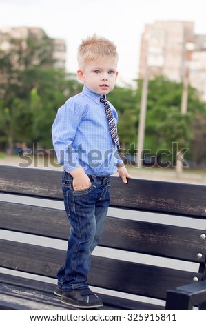 Young boy in the blue shirt, jeans and striped tie is standing on the wooden bench in the park. Outdoor shot