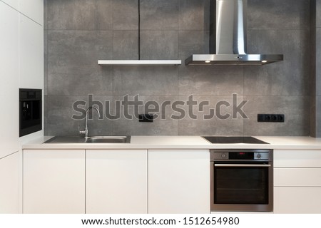Modern interior design at white contemporary kitchen in loft style. Glossy cabinet with built in household appliance, electric stove, oven, sink on worktop and extractor hood on grey wall Сток-фото © 