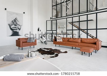 Loft style interior in house with modern living room, bath in bathroom, comfortable couch, pillows on leather armchair, painting on wall and scooter on concrete floor with skin carpet
