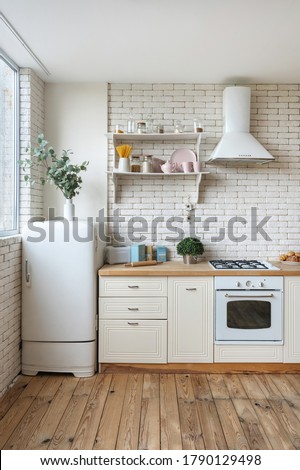 Vertical view of fridge, gas stove appliance, cooking hood, built in oven equipment, kitchenware supplies on shelves and green plants in kitchen at modern house with white interior design