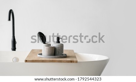 Modern bathroom interior with bathtub and water tap. Panoramic view of tray with hairbrush, soap in bottle dispenser and clean towels at wooden shelf on white contemporary bath near black faucet