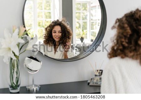 Natural beauty and haircare concept. Charming african american woman in bathrobe looking at mirror in bathroom, touching fresh healthy hair and smiling wide after spa procedure at home