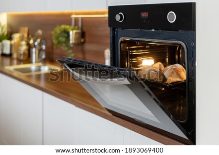 Homemade baking. Open door at electric oven with air ventilation and tray full of whole fresh loaf. Side view of modern technology appliance against kitchen furniture on copy space background