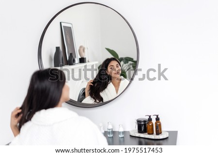 Concept of natural beauty and haircare treatment at home. Young asian woman in bathrobe sitting behind makeup table in white modern bathroom, looking at mirror reflection, touching healthy brown hair.