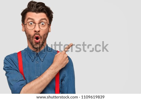 Surprised male hipster, keeps mouth opened, being stupefied as advertises something, wears fashionable shirt and red suspenders, indicates with fore finger at upper right corner. Omg concept Stockfoto © 