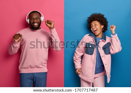 Horizontal shot of ethnic boyfriend and girlfriend dance carefree, enjoy awesome headphones bits, smile happily, raise hands, have fun together at disco party, pose against two colored background