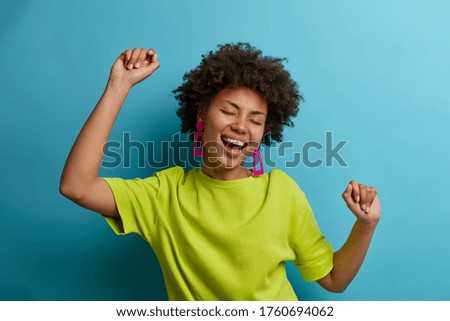 I am winner! Ecstatic overjoyed African American woman dances carefree, celebrates victory and success, dressed in green casual t shirt, feels lively and energetic, isolated on blue background