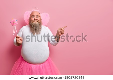 Funny man wears fairy costume, invites you on holiday or costume party, indicates right at blank space, holds magic wand, poses against rosy wall. Dad entertains children during birthday celebration