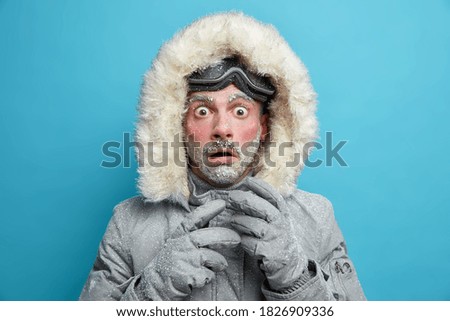 Frozen male explorer has red face covered with frost stares very shocked at camera surprised by very low temperature wears warm jacket and gloves has walk outdoor during blizzard cold weather