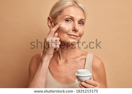 Close up shot of middle aged beautiful woman applies anti aging cream on face undergoes beauty treatments cares about skin poses against beige background. Wrinkled female model with cosmetic product