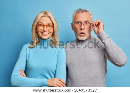 Cheerful middle aged woman and her husband pensioner reacts on shocking news keeps hand on spectacles isolated over blue background. Family age and emotions concept. Senior couple at pose home