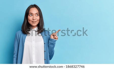 Look at this advert. Pleased brunette young Asian woman points aside shows blank copy space for commercial text promo idea presentation poses against blue background. Attention to nice product