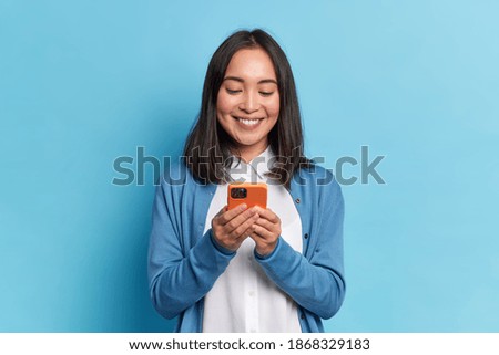 Smiling charming brunette Asian woman uses mobile phone happy texting in social networks addicted to modern technologies wears casual jumper isolated over blue background. Online communication