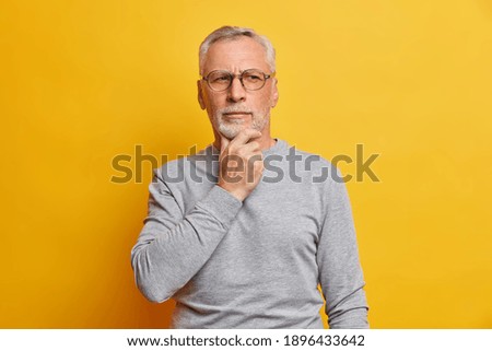 Senior thougthful man holds chin and looks pensively aside makes plannings wears spectacles and casual grey jumper isolated over vivid yellow background. Handsome grandfather considers something
