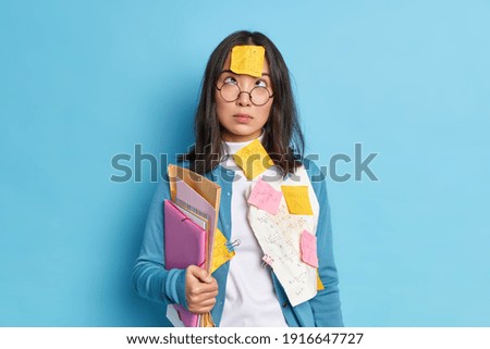 Studio shot of brunette young Asian schoolgirl studies mathematics concentrated above on forehead with sticker holds folders wears round spectacles and sweater isolated over blue background.