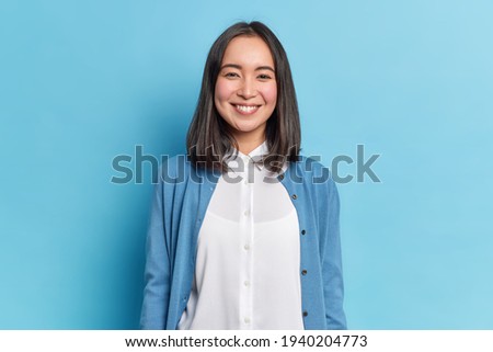 Horizontal shot of pretty Asian woman with dark hair smiles pleasantly looks directly at camera has toothy smile wears white shirt and jumper isolated over blue background. Emotions concept.