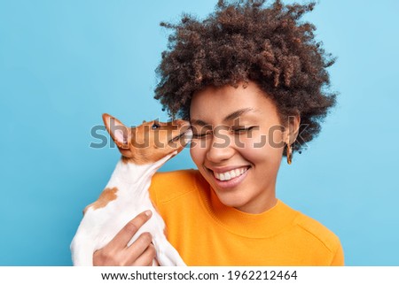 Friend of family. Close up shot of happy curly haired woman plays with dog expresses positive emotions likes animals. Small pedigree puppy licks face of owner. Adopted pet. Tender sincere feelings