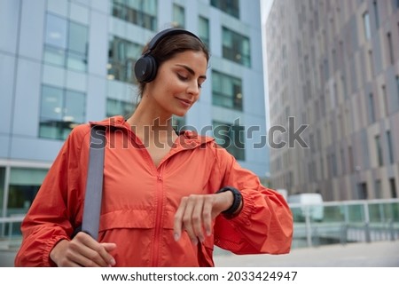 Female runner looks at smartwatch while going for run checks time listens music via headphones dressed in jacket strolls outdoor against modern tall buildings tracks wearable device wanders in city