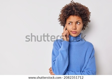 Photo of serious young curly woman keeps finger on temple tries to concentrate considers something contemplates about important thing wears casual blue jumper isolated over white background. ストックフォト © 