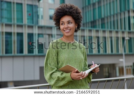 Outdoor shot of thoughtful curly haired woman stands against urban building writes information in notebook holds pen and notepad notes text creats new chapter o book dressed in casual green jumper. Foto stock © 