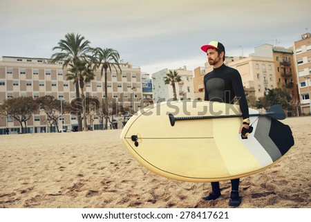 Young attractive surfer sports man wearing a neoprene diving suit and looking at the horizon while carrying his surfing board during a cloudy day